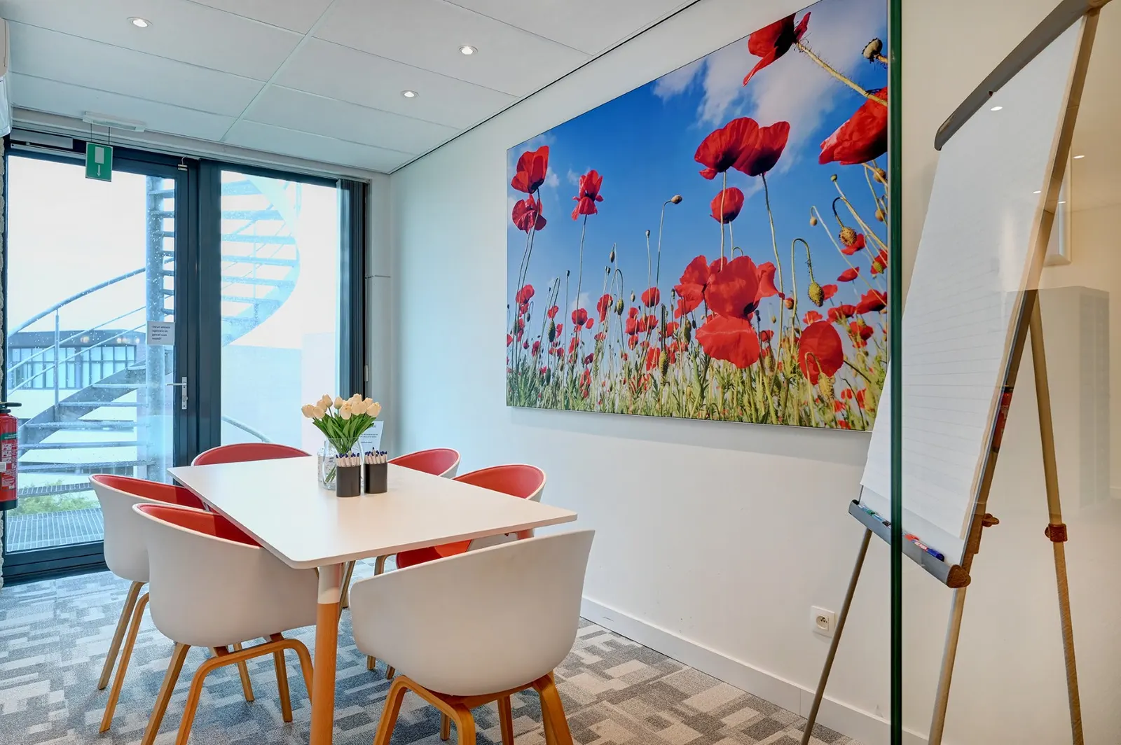 Ronde overlegtafel met witte stoelen, flipchart en een schilderij met klaprozen in Office Capital Hasselt