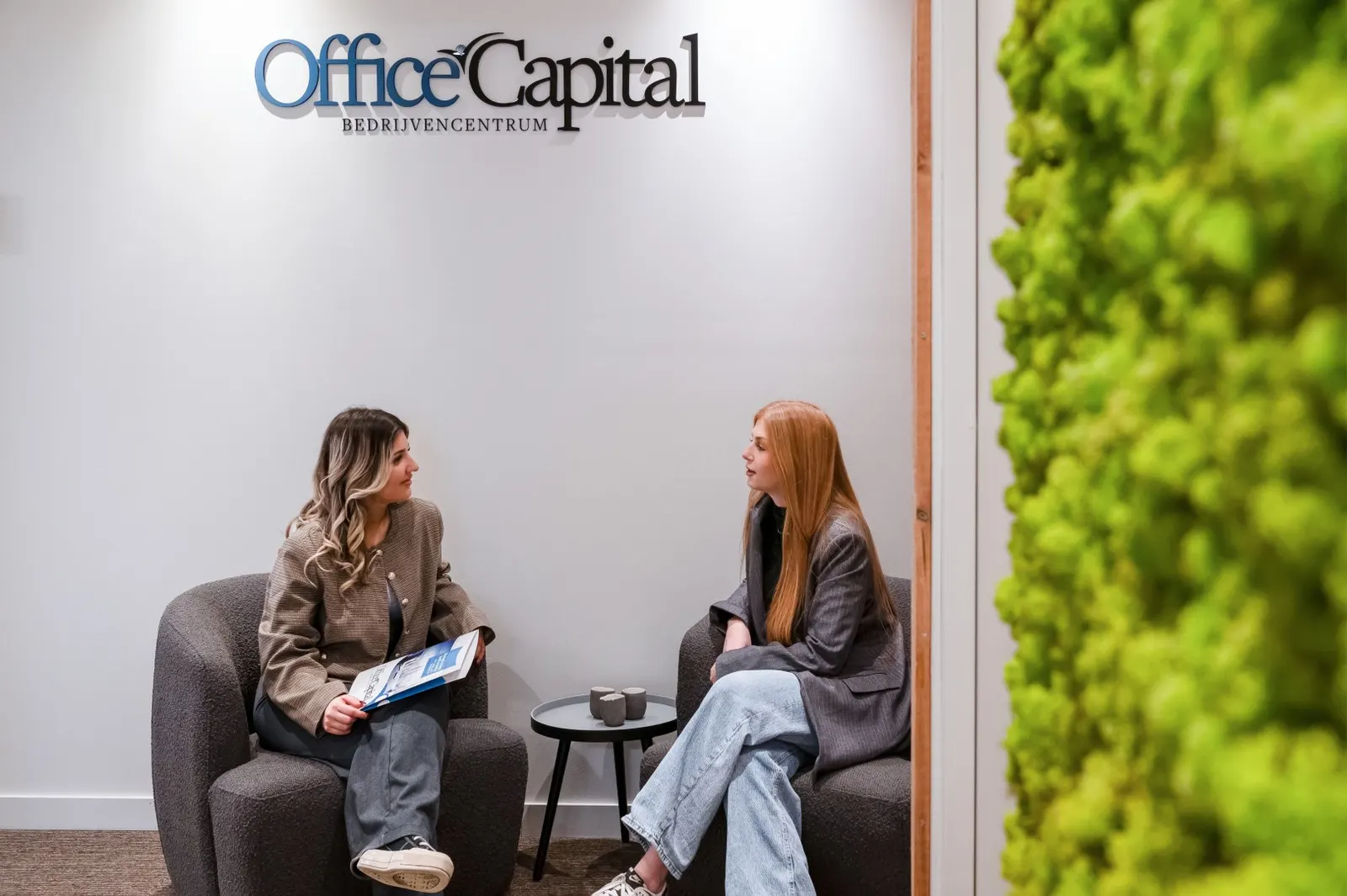 Twee mensen in gesprek in de zithoek bij de groene mosmuur met het logo van Office Capital in Hasselt