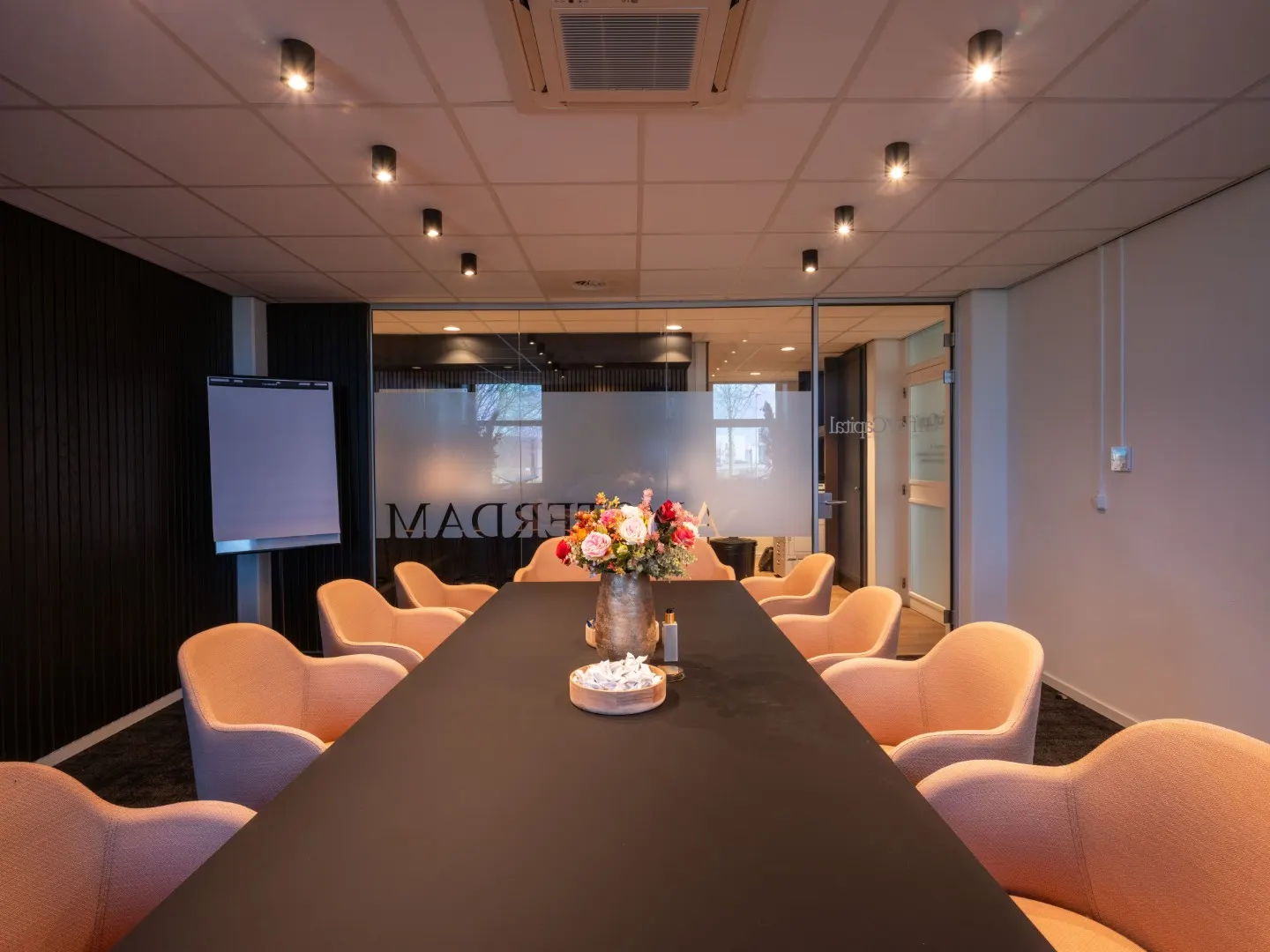 Lange vergadertafel met oranje stoelen en een flipchart in Office Capital Maastricht-Airport